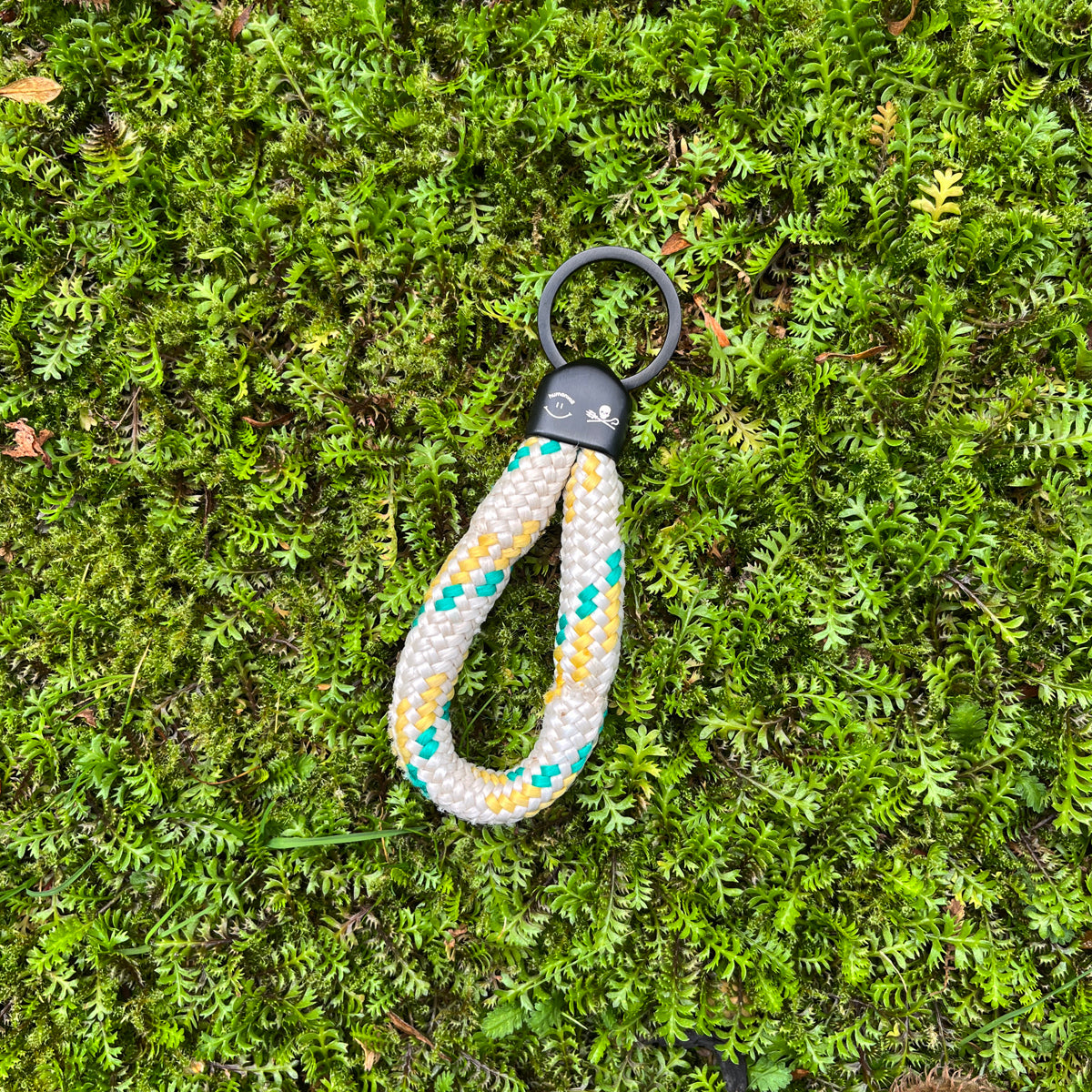 Der Sea Shepherd X BRACENET x humanvoll Schlüsselanhänger mit schwarzer Kappe mit dem Sea Shepherd und humanvoll Logo aus weißem Fischereiseil mit gelben und grünen Akzenten liegt auf grünen Pflanzen