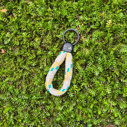 Der Sea Shepherd X BRACENET x humanvoll Schlüsselanhänger mit schwarzer Kappe mit dem Sea Shepherd und humanvoll Logo aus weißem Fischereiseil mit gelben und grünen Akzenten liegt auf grünen Pflanzen