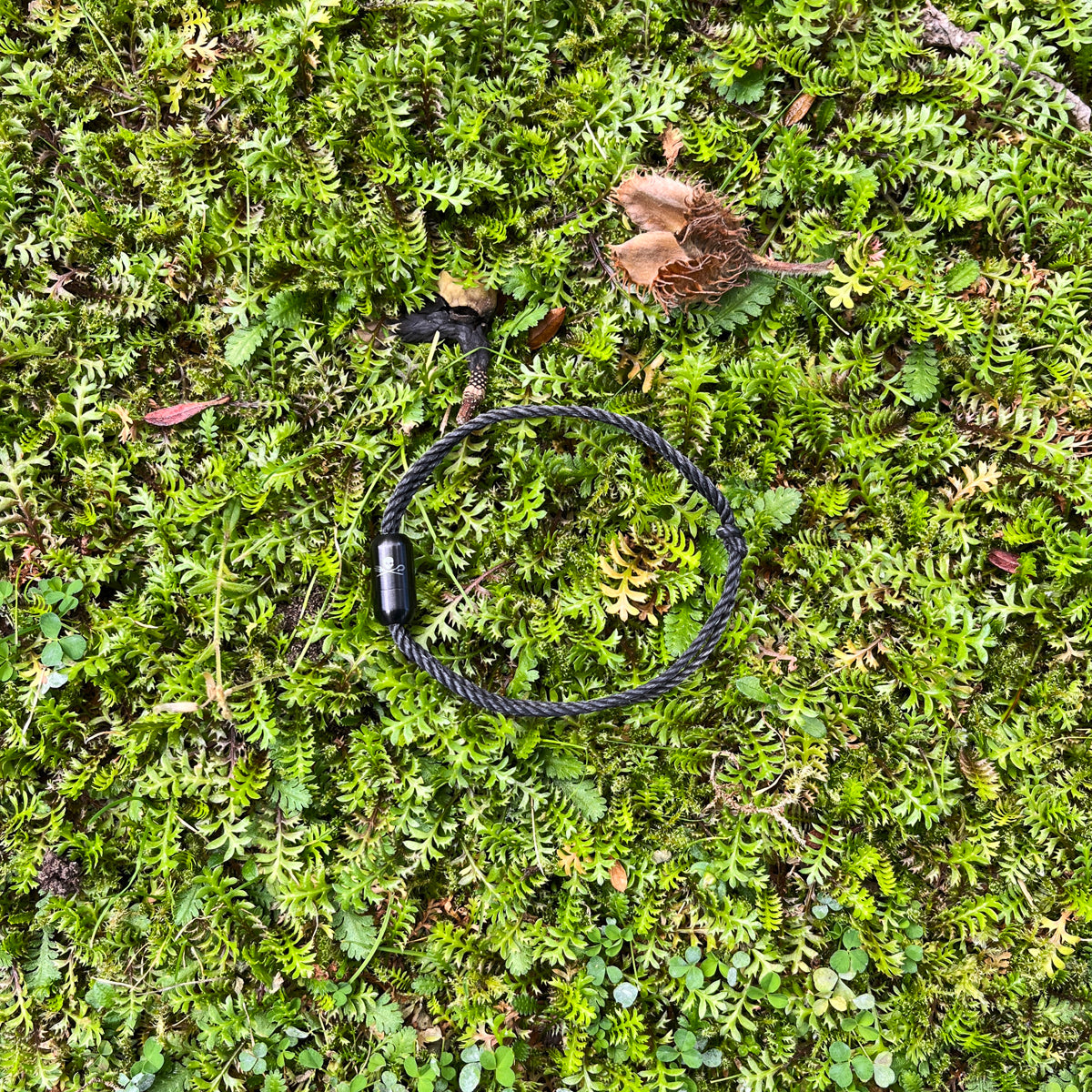 Sea Shepherd X BRACENET x humanvoll Armband aus schwarzen Fischernetzen mit schwarzem Magnetverschluss auf dem das Sea Shepherd Logo, das humanvoll Logo und das BRACENET Logo graviert sind liegt auf grünen Pflanzen