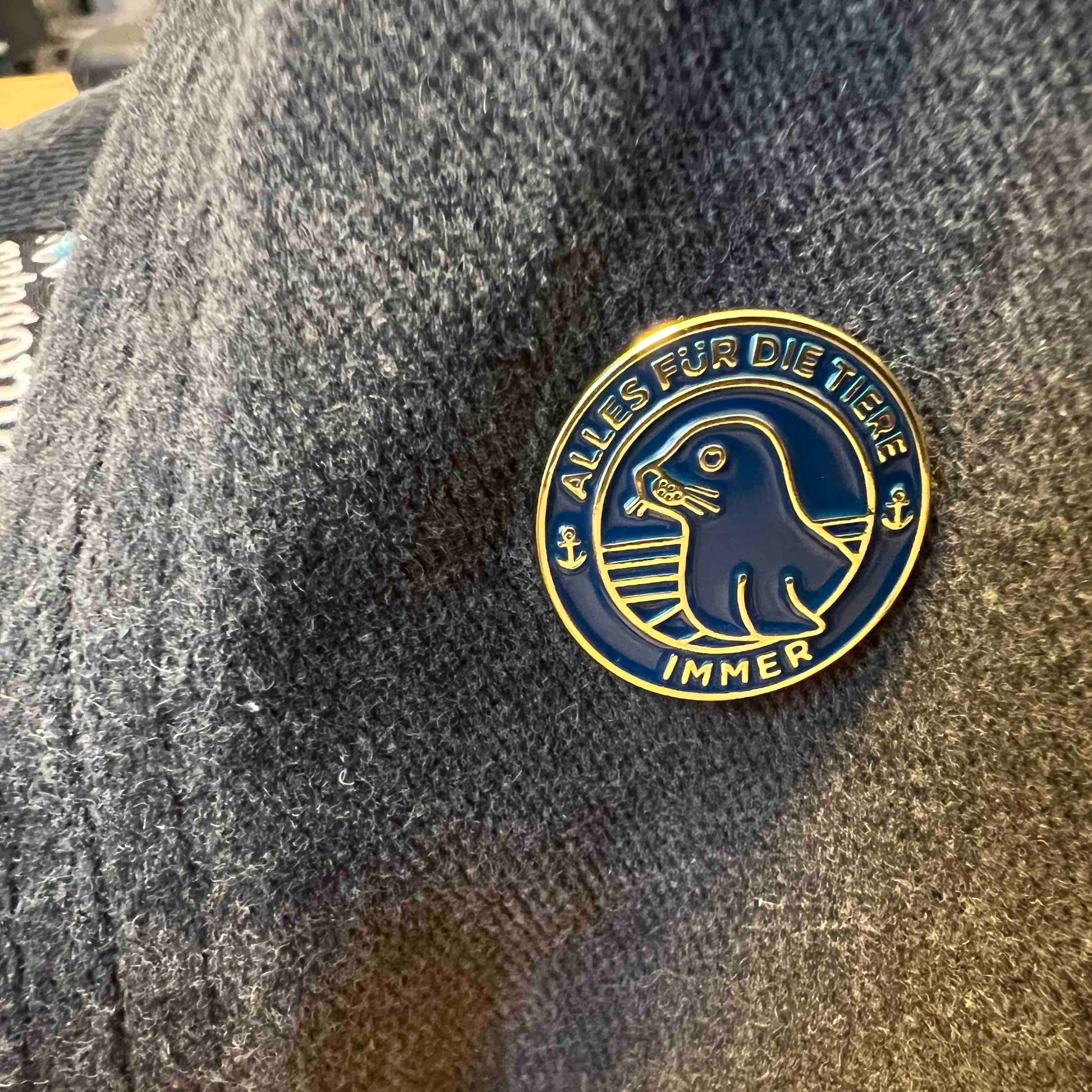 "Alles für die Tiere" Ansteck-Pin aus Emaille in Blau und Gold mit "Alles für die Tiere, immer" Schriftaufzug, sowie Robben-Motiv ist an einer Jacke angebracht.