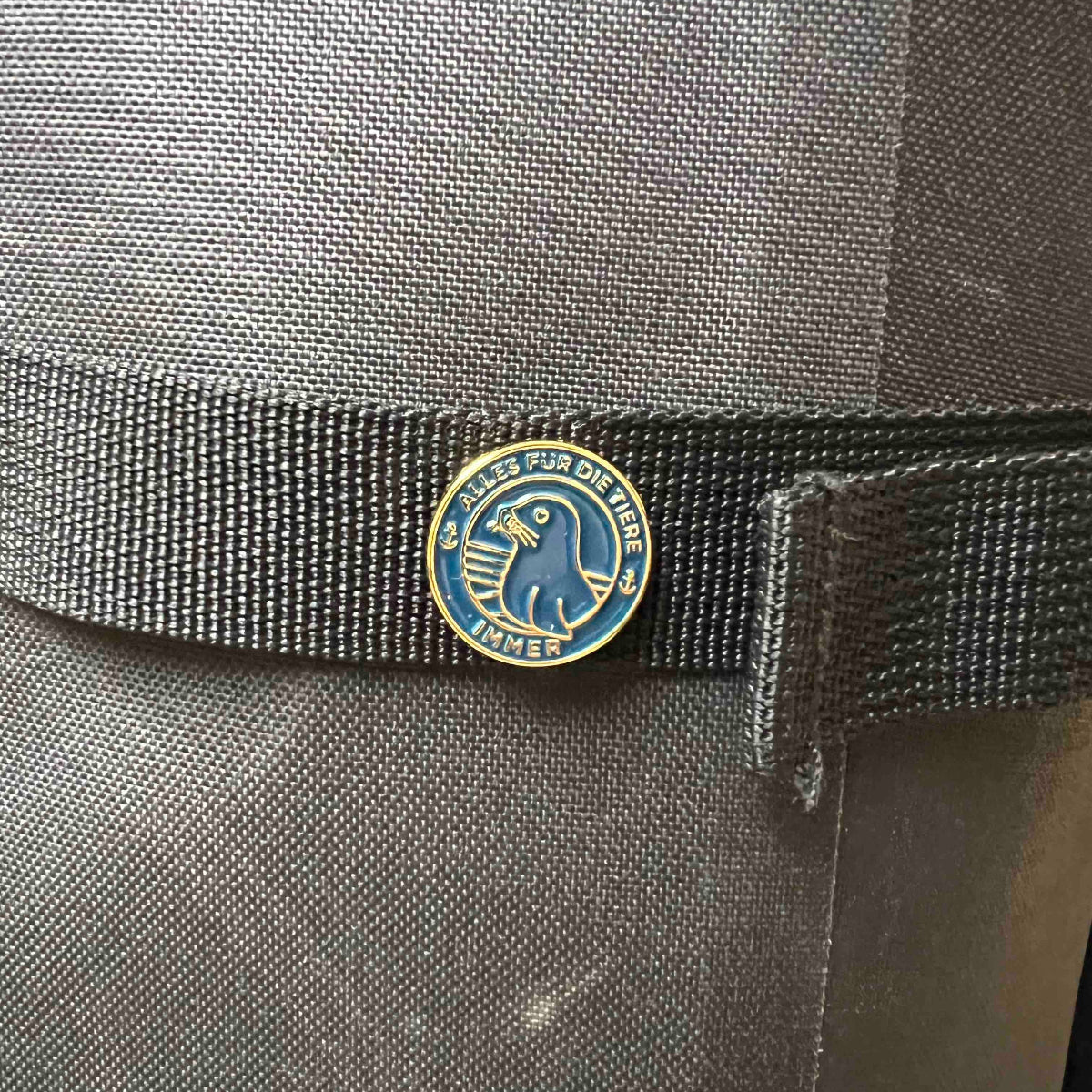 "Alles für die Tiere" Ansteck-Pin aus Emaille in Blau und Gold mit "Alles für die Tiere, immer" Schriftaufzug, sowie Robben-Motiv, ist an einer Tasche angebracht.