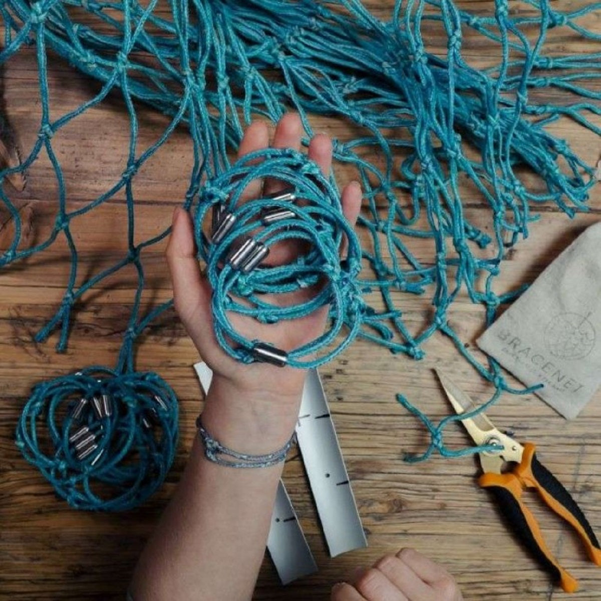 Person hält mehrere handgemachte BRACENET North Sea II Armbänder in der Hand, im Hintergrund Materialien aus der Fertigung