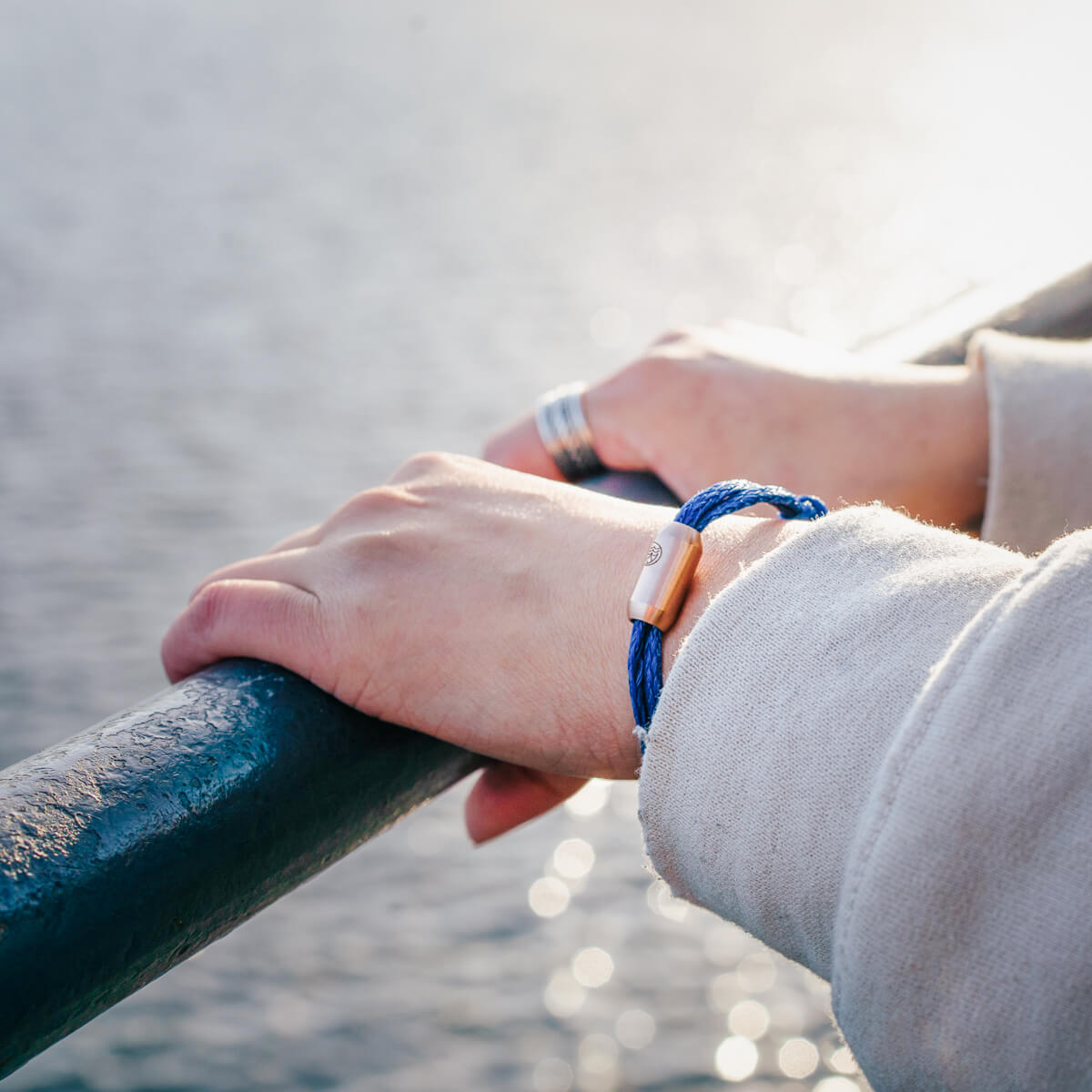 Person trägt das BRACENET Upcycling Armband Baffin Bay aus alten blauen Fischernetzen mit roségoldener Tube am Handgelenk und steht am Ufer.