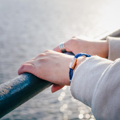 Person trägt das BRACENET Upcycling Armband Baffin Bay aus alten blauen Fischernetzen mit roségoldener Tube am Handgelenk und steht am Ufer.