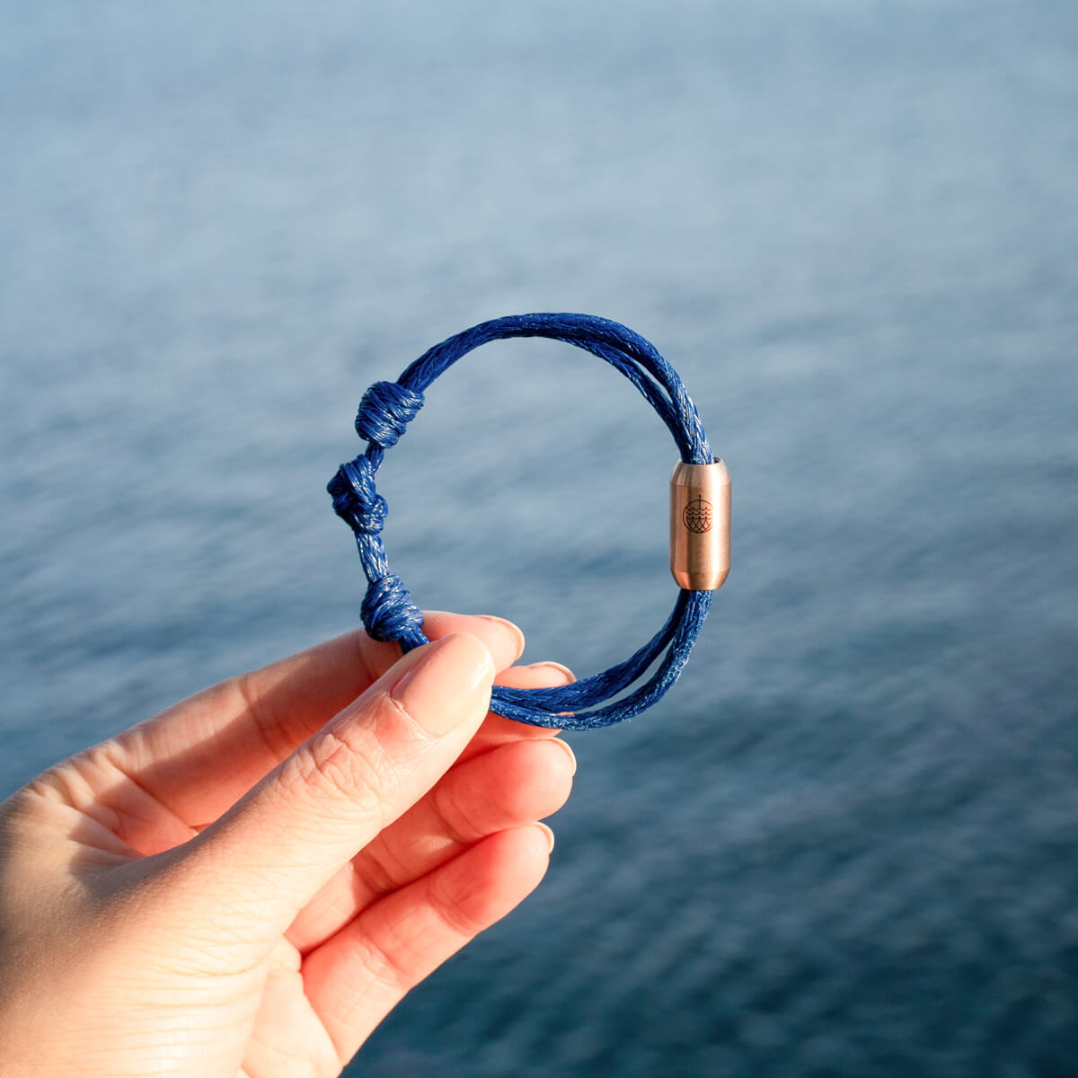 Person hält das BRACENET Upcycling Armband Baffin Bay aus alten blauen Fischernetzen mit roségoldener Tube in der Hand, im Hintergrund das Meer.