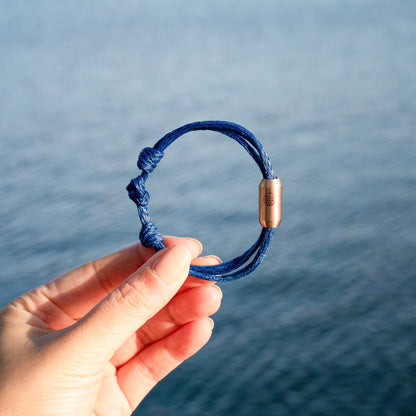 Person hält das BRACENET Upcycling Armband Baffin Bay aus alten blauen Fischernetzen mit roségoldener Tube in der Hand, im Hintergrund das Meer.