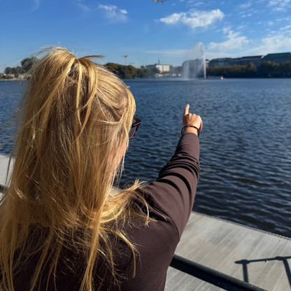 Person trägt das Oktober Bracenet Cardigan Bay aus einem Strang Fischernetz in Schwarz und einem Strang Fischernetz in Orange, verbunden mit einem Knoten und einem Magnetverschluss in Schwarz am Handgelenk und zeigt auf den See. 