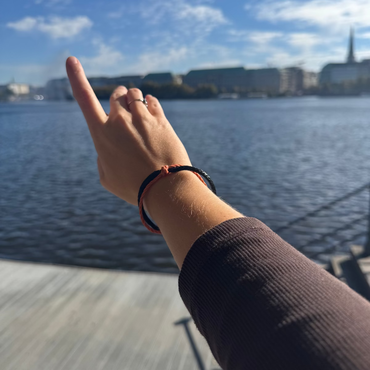 Person trägt das Oktober Bracenet Cardigan Bay aus einem Strang Fischernetz in Schwarz und einem Strang Fischernetz in Orange, verbunden mit einem Knoten und einem Magnetverschluss in Schwarz am Handgelenk und zeigt auf etwas. 