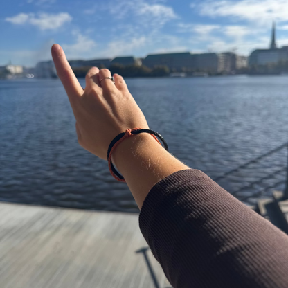 Person trägt das Oktober Bracenet Cardigan Bay aus einem Strang Fischernetz in Schwarz und einem Strang Fischernetz in Orange, verbunden mit einem Knoten und einem Magnetverschluss in Schwarz am Handgelenk und zeigt auf etwas. 