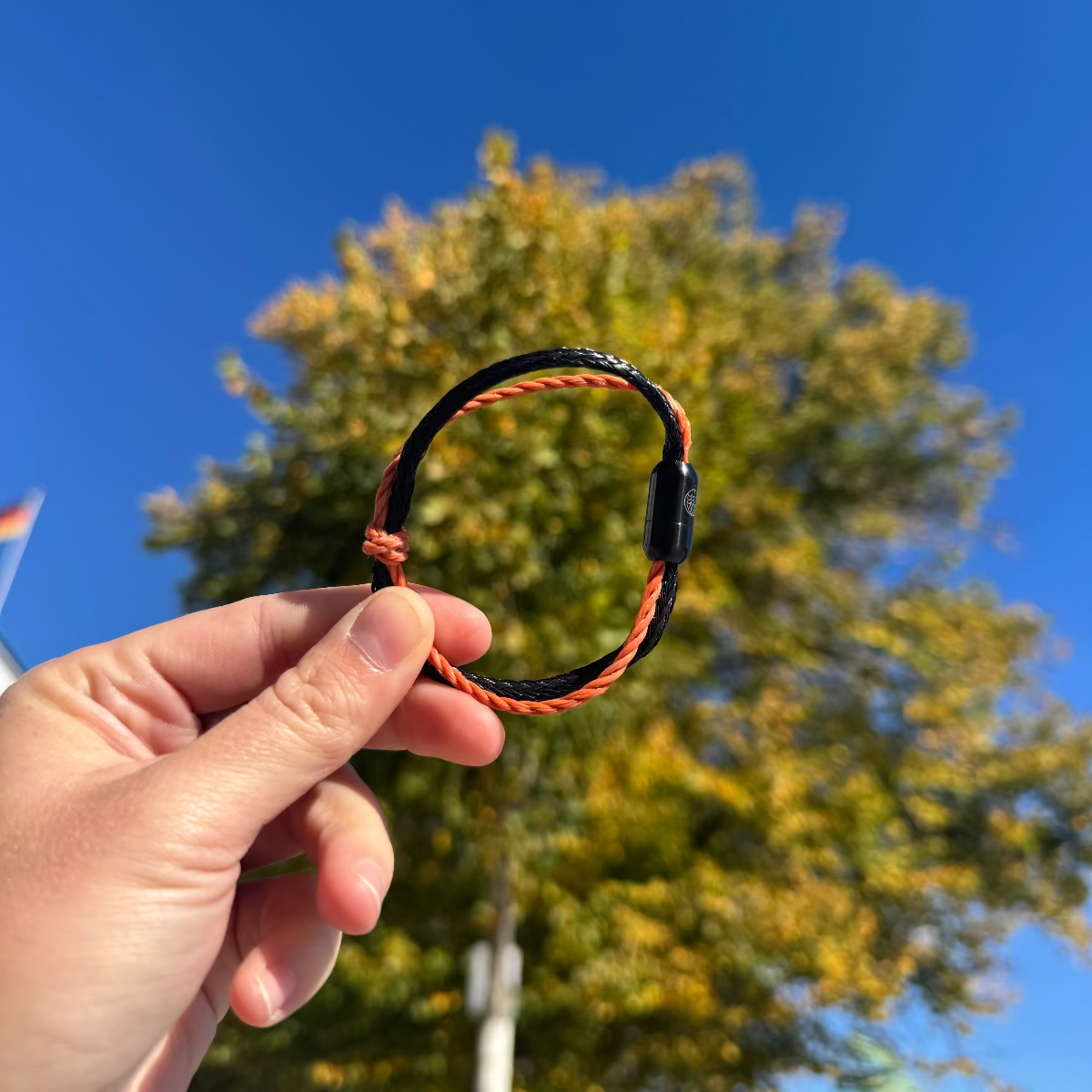Person hält das Oktober Bracenet Cardigan Bay aus einem Strang Fischernetz in Schwarz und einem Strang Fischernetz in Orange, verbunden mit einem Knoten und einem Magnetverschluss in Schwarz zeischern den Fingern. Im Hintergrund ist ein Baum.