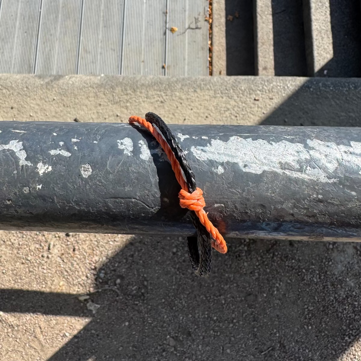 Oktober Bracenet Cardigan Bay aus einem Strang Fischernetz in Schwarz und einem Strang Fischernetz in Orange, verbunden mit einem Knoten und einem Magnetverschluss in Schwarz ist an einem Geländer angebracht. 
