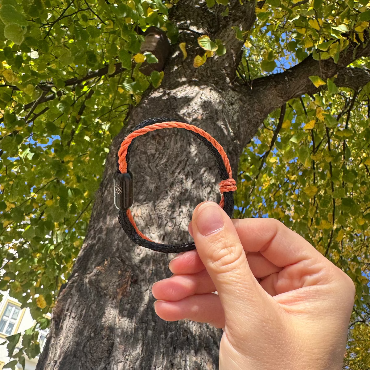 Person hält das Oktober Bracenet Cardigan Bay aus einem Strang Fischernetz in Schwarz und einem Strang Fischernetz in Orange, verbunden mit einem Knoten und einem Magnetverschluss in Schwarz zwischen den Fingern und hält es vor einen Baum.