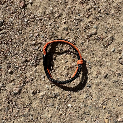 Oktober Bracenet Cardigan Bay aus einem Strang Fischernetz in Schwarz und einem Strang Fischernetz in Orange, verbunden mit einem Knoten und einem Magnetverschluss in Schwarz liegt auf dem Boden.