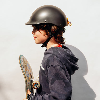 Junge trägt Dashel x BRACENET Kids' Sea Shepherd Fahrradhelm in Schwarz mit Jolly Roger Logo und Fischernetz-Schlaufe in Gelb und Blau und hält ein Skateboard.