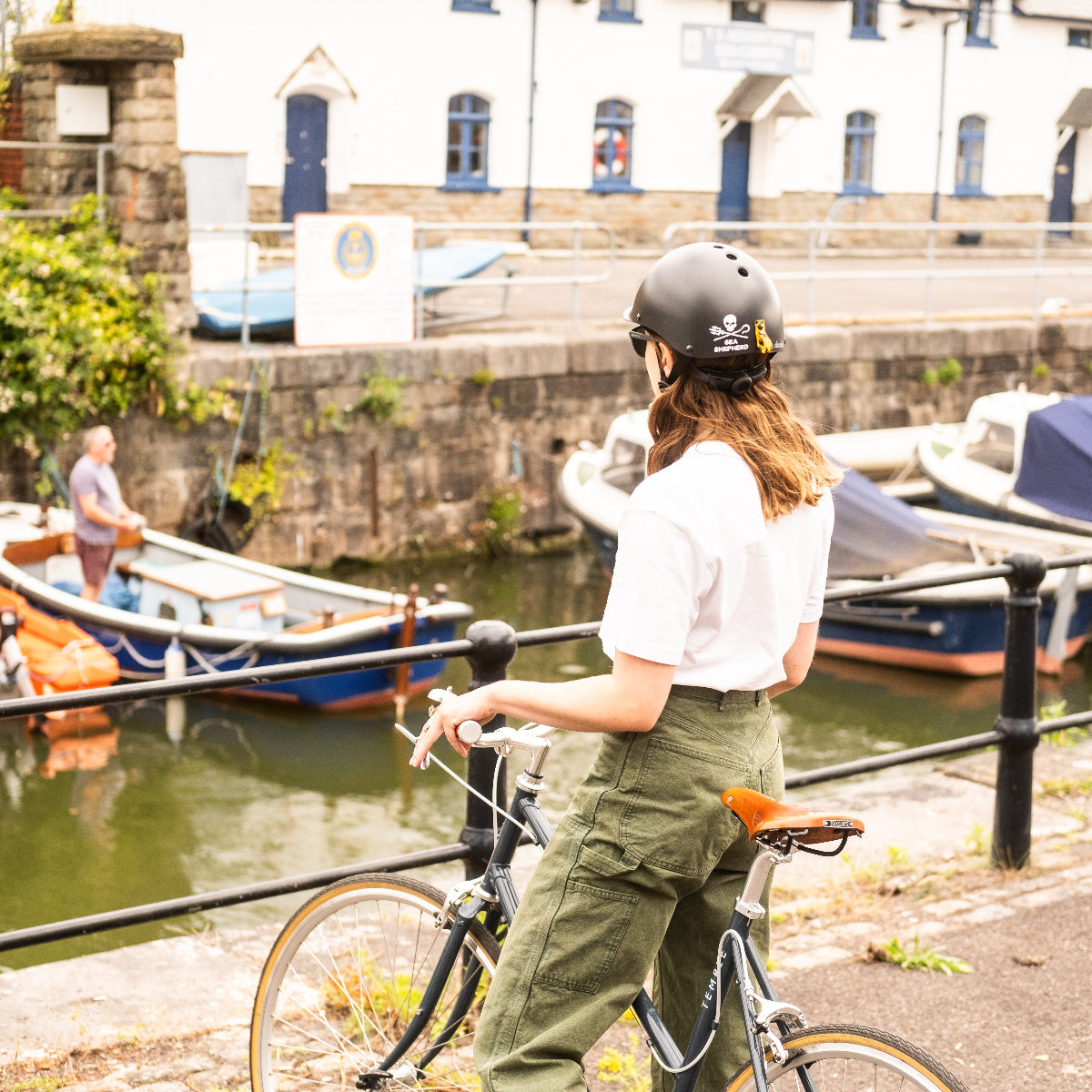 Person trägt Dashel x BRACENET Sea Shepherd Fahrradhelm in Schwarz mit Sea Shepherd Logo und Fischernetz-Schlaufe in Gelb und Blau und steht am Fluss. 