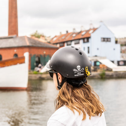 Person trägt Dashel x BRACENET Sea Shepherd Fahrradhelm in Schwarz mit Sea Shepherd Logo und Fischernetz-Schlaufe in Gelb und Blau und schaut auf den Fluss. 