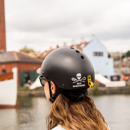 Person trägt Dashel x BRACENET Sea Shepherd Fahrradhelm in Schwarz mit Sea Shepherd Logo und Fischernetz-Schlaufe in Gelb und Blau und schaut auf einen Fluss. 