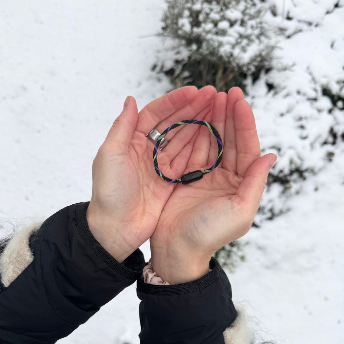 Person hält das Januar Bracenet Kara Sea aus schwarzem Fischernetz mit Umwicklungen aus Dolly Ropes in Grün und Lila mit schwarzem Bajonettverschluss in den Händen über dem Schnee. 