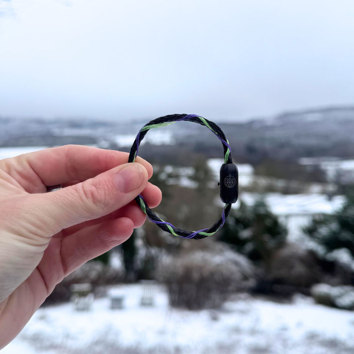 Person hält das Januar Bracenet Kara Sea aus schwarzem Fischernetz mit Umwicklungen aus Dolly Ropes in Grün und Lila mit schwarzem Bajonettverschluss zwischen den Fingen und hält es vor die verschneite Landschaft.