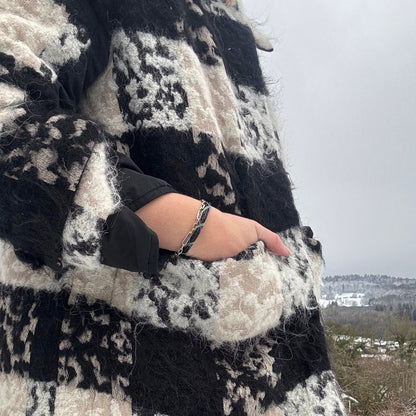 Person trägt das Januar Bracenet Kara Sea aus schwarzem Fischernetz mit Umwicklungen aus Dolly Ropes in Grün und Lila mit schwarzem Bajonettverschluss Am Handgelenk und hat eine Hand in der Tasche des Mantels. 