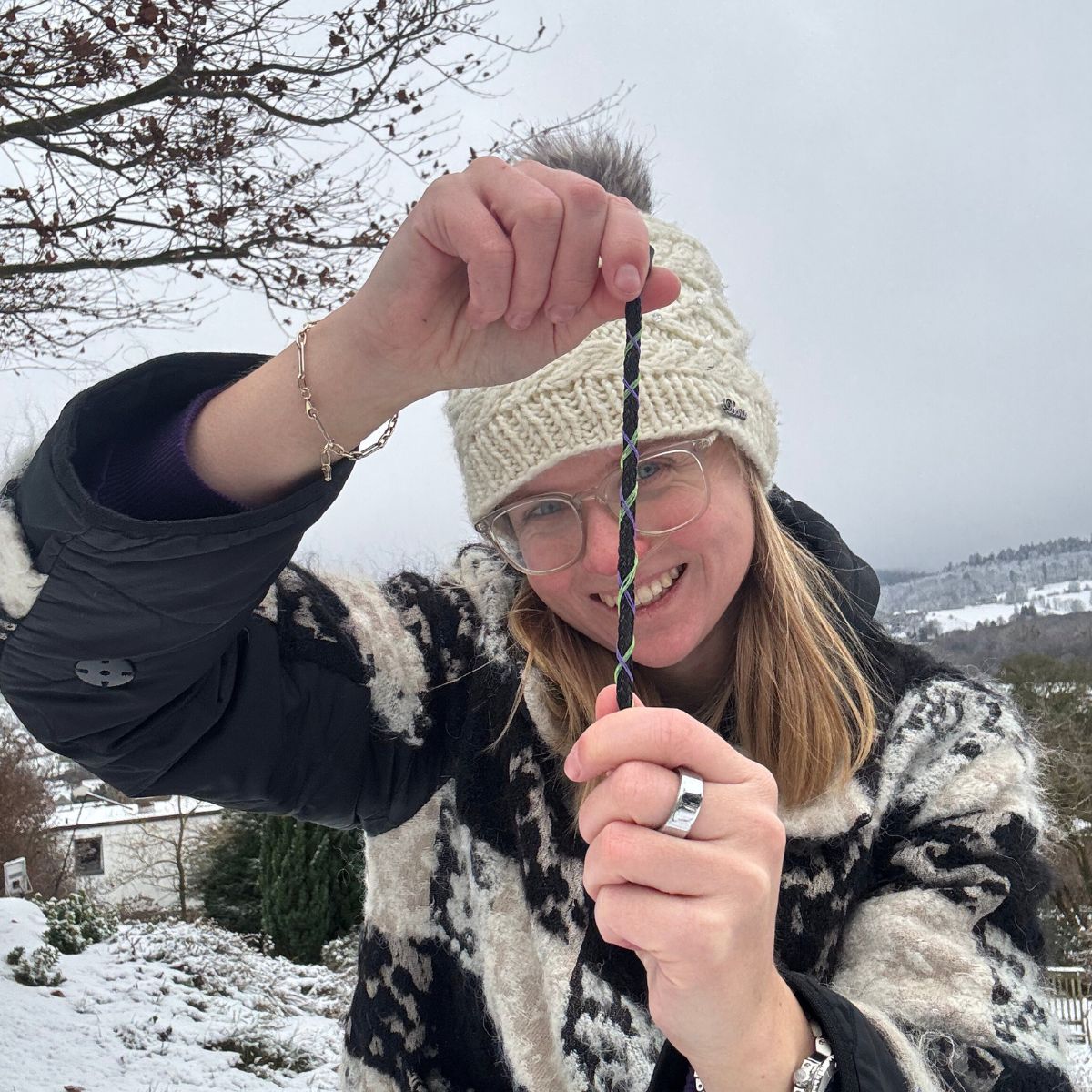 Person hält das Januar Bracenet Kara Sea aus schwarzem Fischernetz mit Umwicklungen aus Dolly Ropes in Grün und Lila mit schwarzem Bajonettverschluss gestreckt vor das Gesicht und lacht. Es liegt Schnee. 