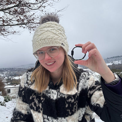 Person hält das Januar Bracenet Kara Sea aus schwarzem Fischernetz mit Umwicklungen aus Dolly Ropes in Grün und Lila mit schwarzem Bajonettverschluss zwischen zwei Fingern und hält es sich neben das Gesicht. Es liegt Schnee. 