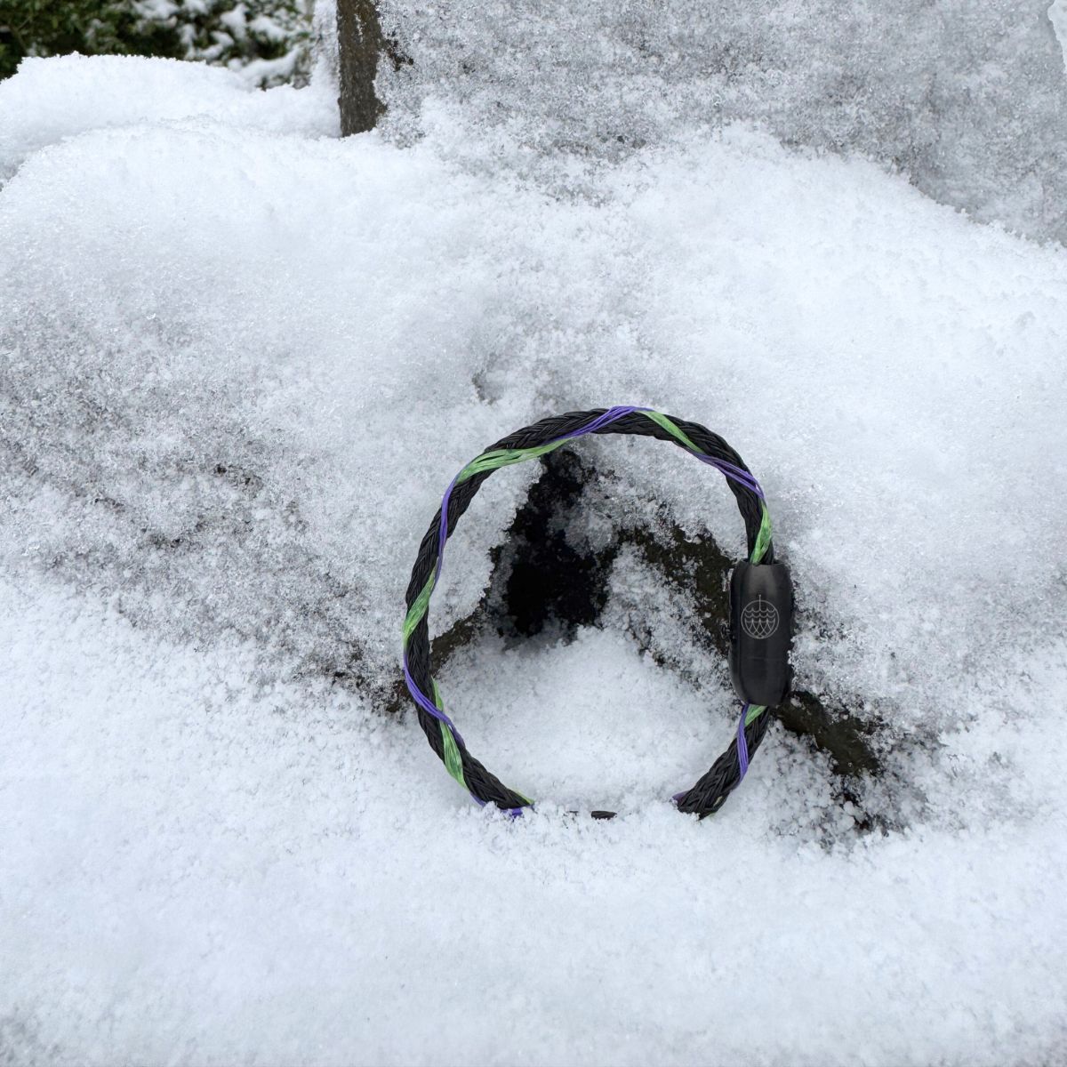 Das Januar Bracenet Kara Sea aus schwarzem Fischernetz mit Umwicklungen aus Dolly Ropes in Grün und Lila mit schwarzem Bajonettverschluss steht im Schnee an einen Stein gelehnt. 