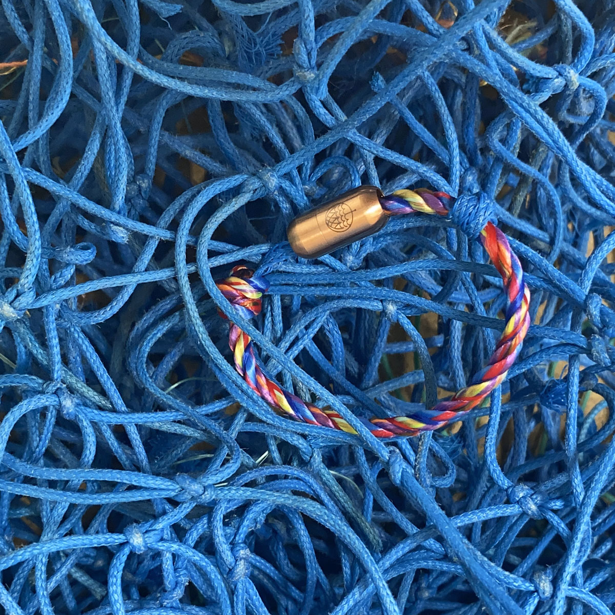 Kids' Bracenet Lübecker Bucht aus einem Strang blauem Fischernetz und einem Strang buntem Fischernetz, größenverstellbar mit Magnetverschluss in Roségold liegt auf Fischernetzen. 