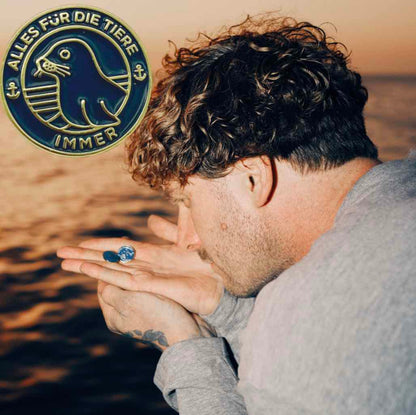 "Alles für die Tiere" Ansteck-Pin aus Emaille in Blau und Gold mit "Alles für die Tiere, immer" Schriftaufzug, sowie Robben-Motiv. Im Hintergrund ist Malte Zierden mit den Pins an der Hand am Meer zu sehen. 