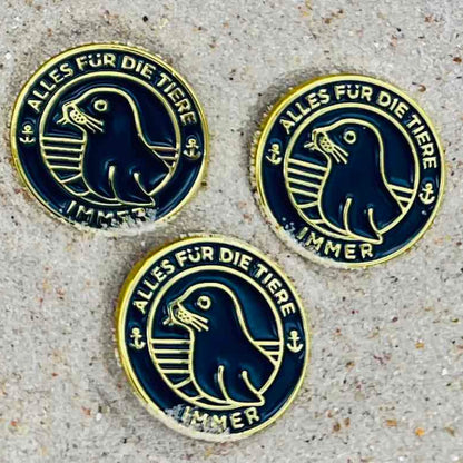 Drei "Alles für die Tiere" Ansteck-Pins aus Emaille in Blau und Gold mit "Alles für die Tiere, immer" Schriftaufzug, sowie Robben-Motiv liegen im Sand.