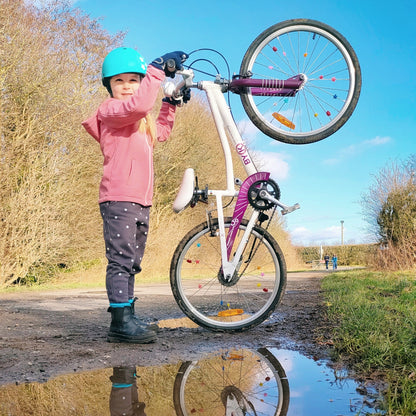 Mädchen trägt Dashel x BRACENET Kids' Ocean Fahrradhelm in Türkis mit Schlaufe aus Fischernetzen in Gelb und Blau und hält ein Fahrrad.