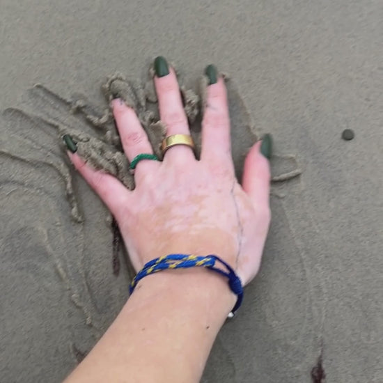 Person trägt das BRACENET Upcycling Armband Insel Sylt II aus alten Fischernetzen in dunkelblau und blau-gelb, mit einem silbernen Magnetverschluss und Anhänger mit einer Inselgravur von Sylt am Handgelenk und streicht durch den Sand. 