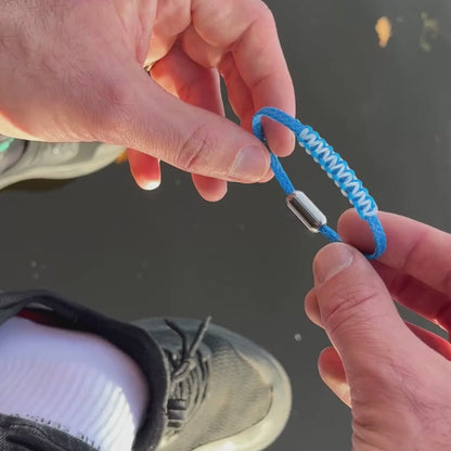Person hält das Dezember BRACENET Upcycling Armband Arctic Ocean II aus blauen Fischernetzen mit Makramee Knoten in Hellblau und Magnetverschluss aus Edelstahl in Silber in den Händen über dem Wasser und dreht das Armband. 