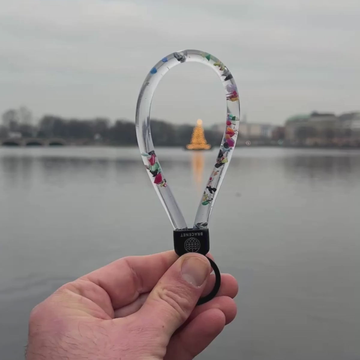 Der BRACENET Floating Upcycling Schlüsselanhänger aus alten Plastiktuben gefüllt mit Mikroplastik, destilliertem Wasser und Glycerin mit Edelstahlkappe und Ring in schwarz wird in das Bild gehalten. Im Hintergrund sieht man einen beleuchteten Weihnachtsbaum. Auf dem Video sieht man wie sich die Mikroplastik-Partikel bewegen. 