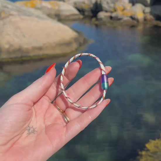 Person zeigt in einem Video das BRACENET Upcycling Armband Rainbow Bay aus alten weißen Fischernetzen, bunt umwickelt, im Hintergrund ein Fjord